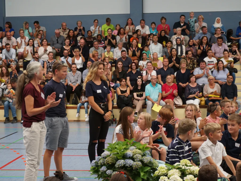 Personen begrüßen die neuen Kinder, das Publikum applaudiert.